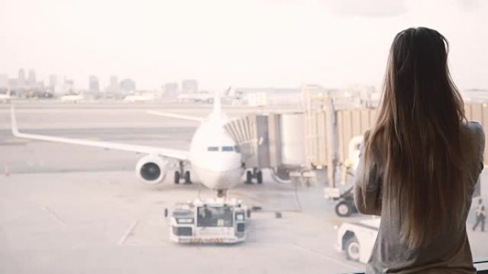 美丽的白人女孩长头发走到机场候机楼的窗口，等待，飞机在背景