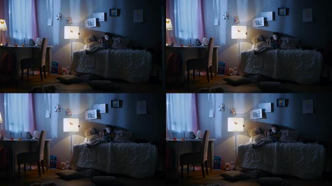 可爱的小女孩躺在床上，在智能手机上看动画片。她的小猫在她旁边玩耍。她的落地灯亮着。