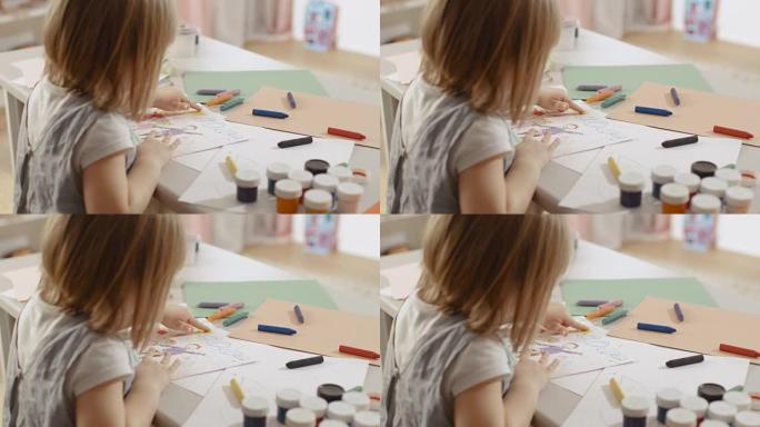 可爱的小女孩坐在她的桌子旁画画。她用黄色手指画太阳。她的房间是粉红色的，舒适的。