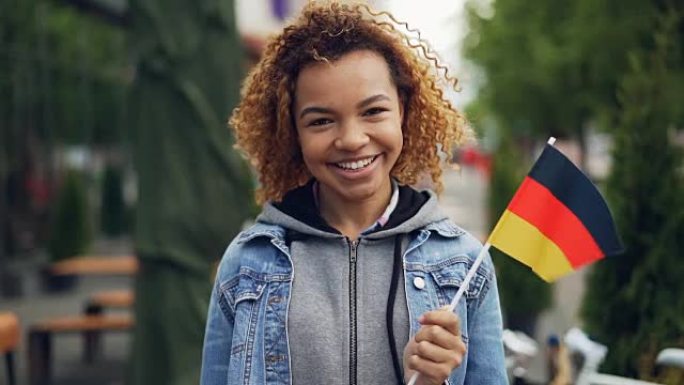 快乐的非洲裔美国女人微笑着的慢动作肖像，举着德国国旗，看着相机，鲜艳的旗帜在风中飘扬。