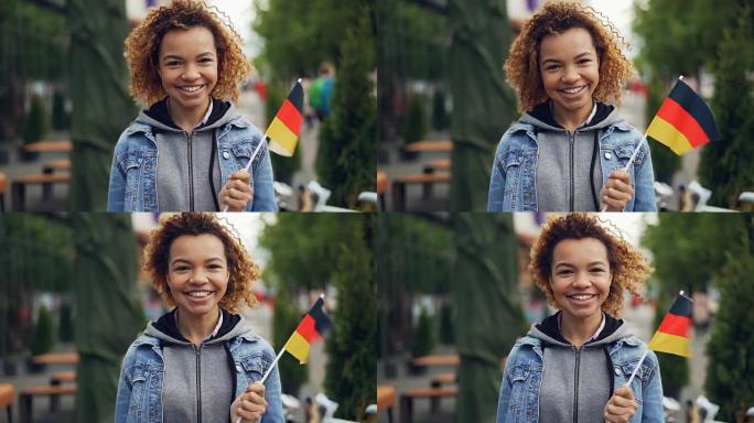 快乐的非洲裔美国女人微笑着的慢动作肖像，举着德国国旗，看着相机，鲜艳的旗帜在风中飘扬。