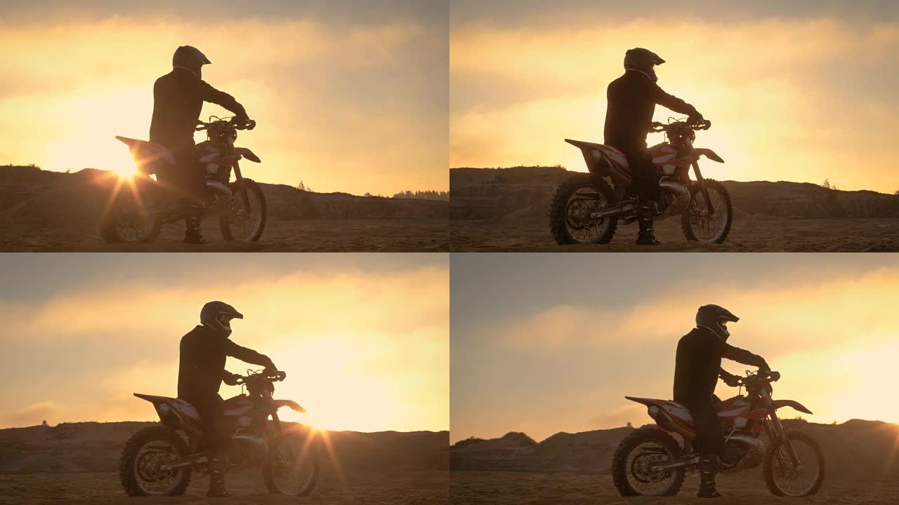专业FMX摩托车骑手在他的摩托车上休息，俯瞰坚硬的沙地越野地形。太阳设置。