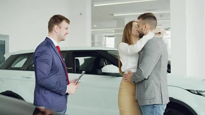 开朗的汽车经销商推销员正在与年轻的买家英俊的男人握手，然后将钥匙扣给他幸福的妻子，夫妻俩正在拥抱和亲