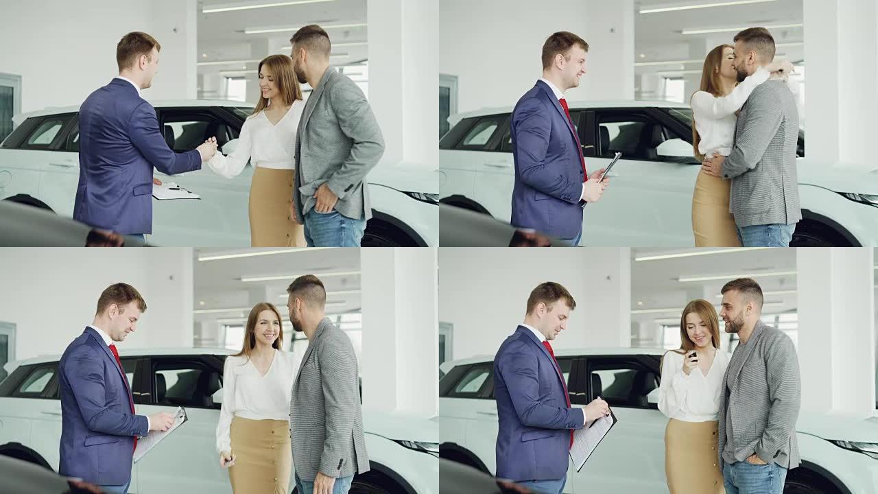 开朗的汽车经销商推销员正在与年轻的买家英俊的男人握手，然后将钥匙扣给他幸福的妻子，夫妻俩正在拥抱和亲