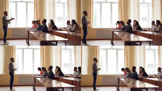 友善的老师正在与学生交谈，聪明的混血人在同学写作时坐在演讲厅的桌子旁举手讲话。