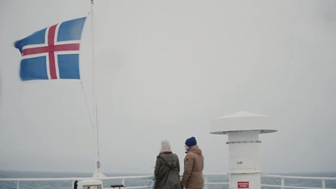 年轻夫妇站在船上，靠近冰岛国旗，看着大海，说话