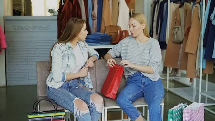 两名年轻女士坐在购物中心的服装部，购物后聊天。背景丰富的女装和购物袋。