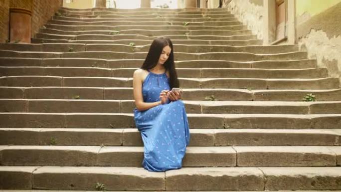 迷人的年轻黑发女人穿着浅色夏装坐在欧洲小镇的楼梯上。她正在看着手机并使用它。