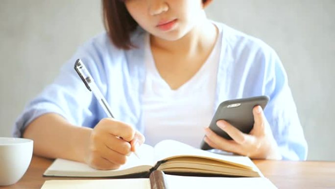 穿着白色连衣裙的年轻女商人坐在咖啡馆的桌子旁，在笔记本上写字。亚洲女人会说智能手机和一杯咖啡。在咖啡