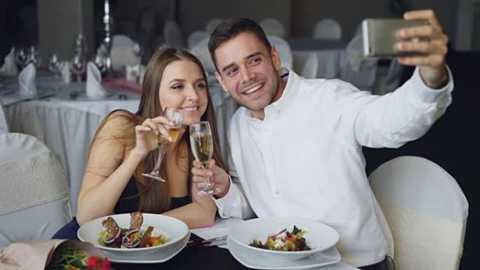 有吸引力的一对情侣在餐厅吃晚饭时，用智能手机用香槟酒杯自拍。他们微笑，亲吻和摆姿势看着相机。