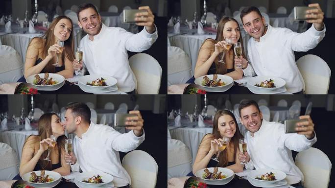 有吸引力的一对情侣在餐厅吃晚饭时，用智能手机用香槟酒杯自拍。他们微笑，亲吻和摆姿势看着相机。