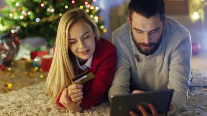 幸福的夫妻躺在圣诞树下的地毯上，女人拿着信用卡，男人在平板电脑上买礼物。