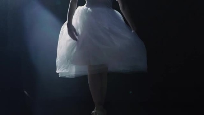 芭蕾舞女演员腿的特写镜头。她在聚光灯下跳舞，穿着尖头芭蕾舞鞋旋转，周围黑暗。她穿着白色短裙。慢动作。