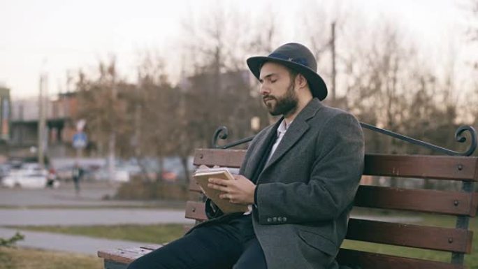 戴着帽子和外套的富有创造力的年轻作家在公园的城市街道长椅上，用笔记本上的笔为他未来的书写笔记