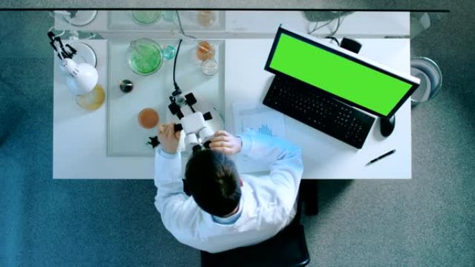 一位科学家的俯视图，该科学家在带有绿色屏幕的个人计算机上工作，看着显微镜并在带有图表的文档上写下结果