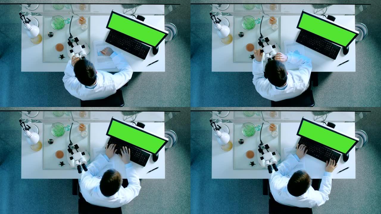 一位科学家的俯视图，该科学家在带有绿色屏幕的个人计算机上工作，看着显微镜并在带有图表的文档上写下结果