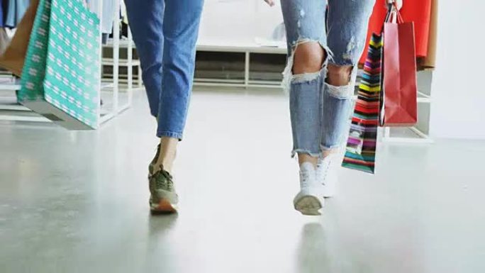 女孩的腿在宽敞的商店里缓慢行走。女士们穿着牛仔裤和现代运动鞋，提着五颜六色的纸袋。