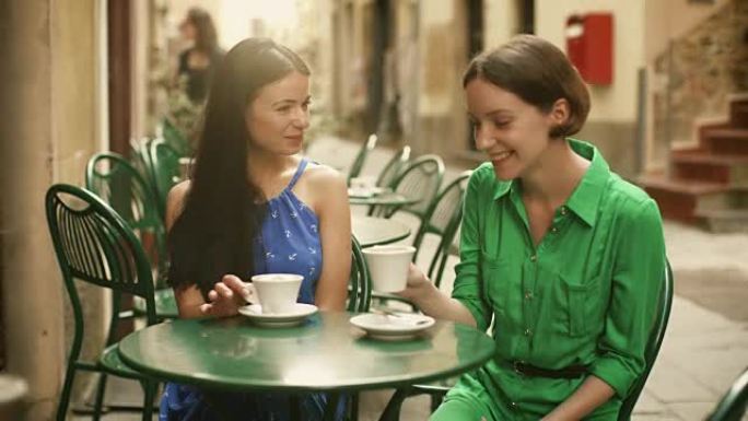 两个穿着浅色夏装的迷人年轻女性在街头咖啡店放松和交谈。女人心情很好，面带微笑。