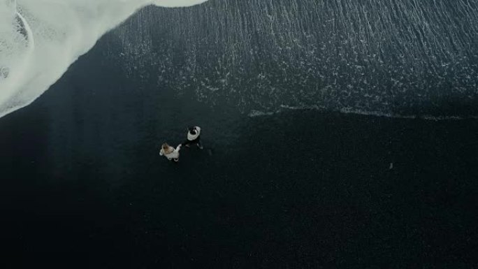 这对年轻夫妇站在冰岛黑海滩的海边欣赏水的空中俯视图