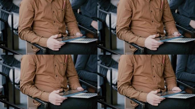 坐在椅子上的笔记本和数字平板电脑的年轻人的特写镜头。大学研讨会或讲座中的男性