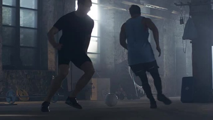 两个肌肉发达的足球运动员在工业体育馆里玩球。他们玩得很开心，各种健身器材躺在地板上。