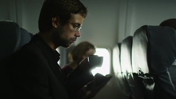 年轻人在飞机上喝咖啡，一个女人在窗户旁边的背景中看书。
