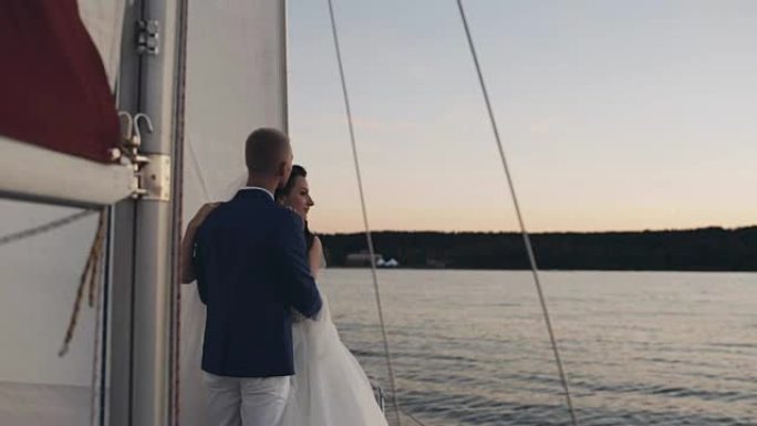 美丽的新婚夫妇拥抱，站在帆船上。穿着婚纱的新娘和新郎看着远处