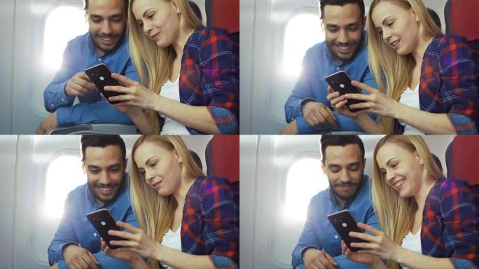 在商用飞机上，漂亮的年轻金发女郎与英俊的西班牙裔男性在智能手机上观看社交媒体并大笑。阳光透过飞机的窗