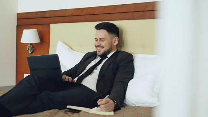 微笑的商人躺在床上，使用笔记本电脑，在酒店房间工作时在记事本上写笔记。旅游、商务和人的概念