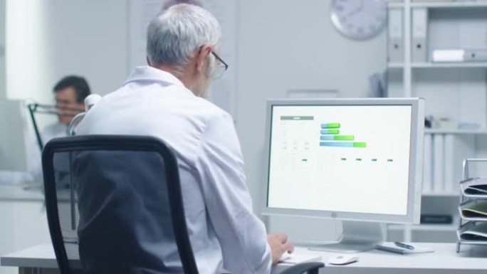高级男性实验室研究员在他的个人电脑上处理图表。他的助手在他的办公桌前工作。