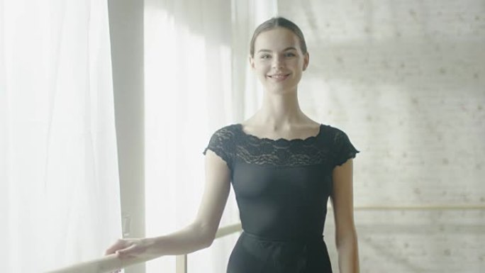美丽的年轻芭蕾舞演员微笑的肖像。在背景中芭蕾舞演员练习。在阳光明媚的现代工作室拍摄。慢动作。