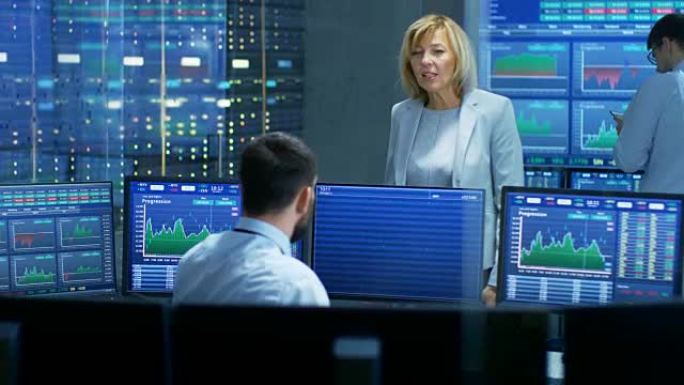 在股市买卖部门，女经理与男交易员交谈。在后台工作的人们中，屏幕显示了股票代码和图表。