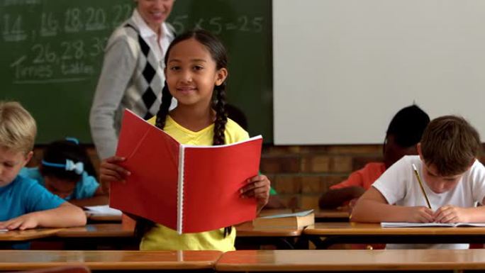 小女孩拿着红色记事本在教室里对着镜头微笑