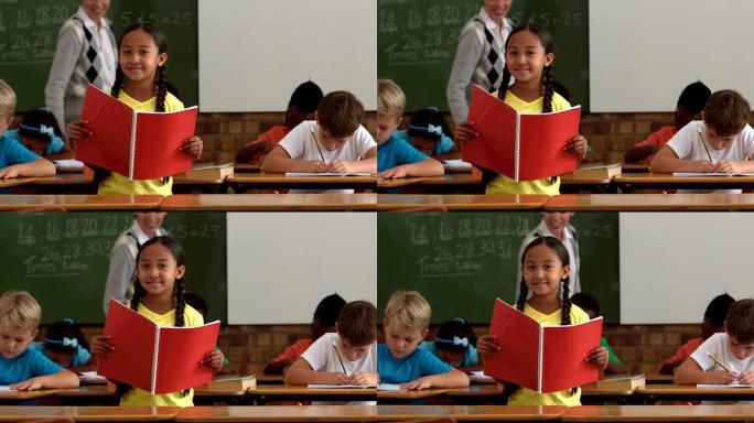 小女孩拿着红色记事本在教室里对着镜头微笑