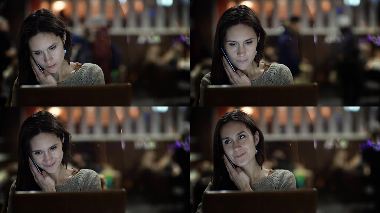 年轻忙碌的女人在咖啡馆打电话的肖像。美女坐在桌旁使用笔记本电脑