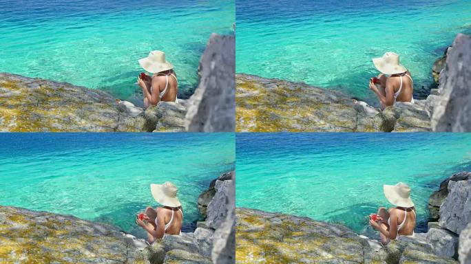 穿着泳衣和太阳帽的女人在宁静的蓝色海洋上吃新鲜的西瓜