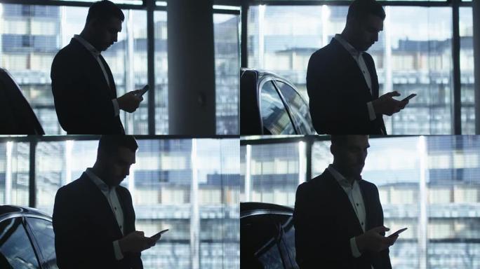 穿着西装的商人在行政轿车旁边使用智能手机，并在车库的大窗户旁使用。