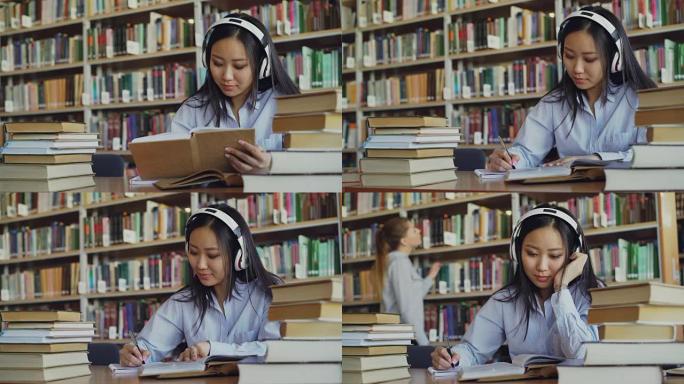 戴着耳机听音乐的亚洲青少年学生正坐在大图书馆的桌子旁，拿着书并写下摘要