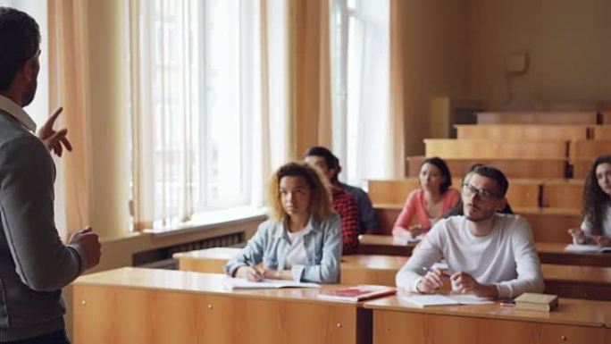 大胡子的教授正在阅读演讲和打手势，而学生则坐在宽敞的大学教室里的桌子旁听和写。
