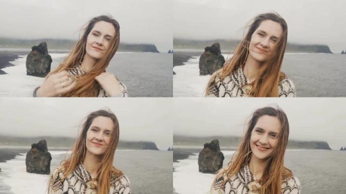 年轻幸福的女人的肖像站在黑海滩上，看着相机，微笑着，靠近冰岛的巨魔脚趾岩石