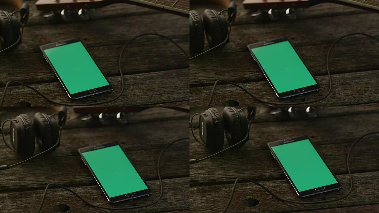 带有绿色屏幕的安卓手机，纵向模式，放在吉他和耳机旁边的木桌上。因果生活方式