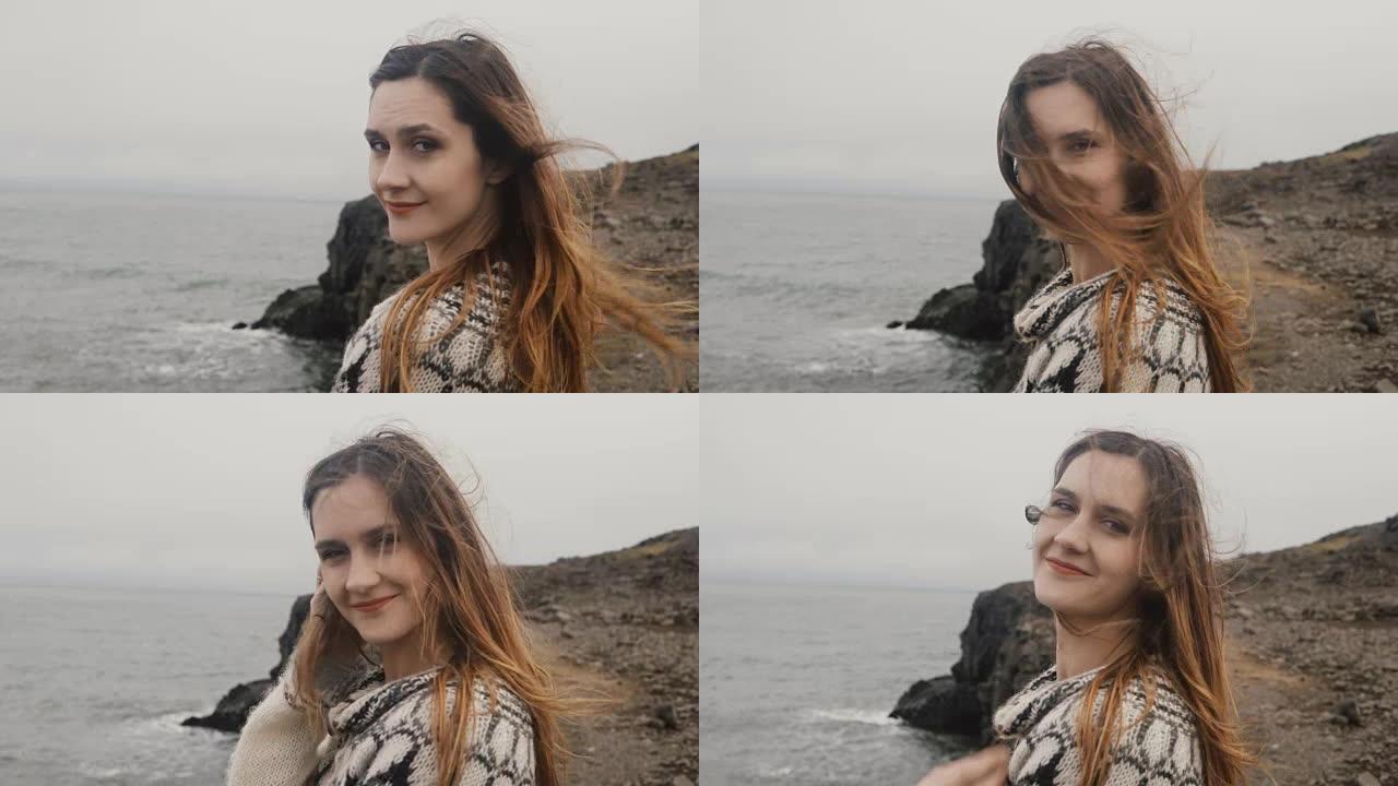 年轻幸福的女人站在海边，看着镜头，微笑着，头发随风飘动的肖像