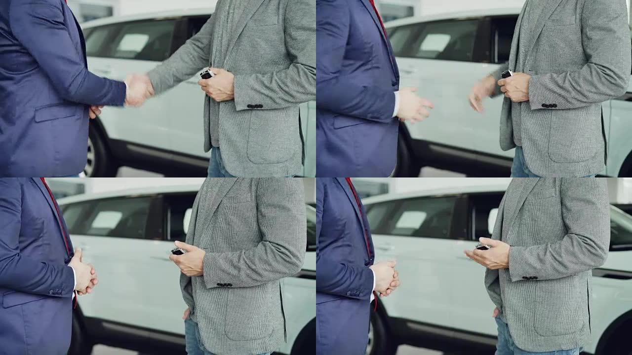 汽车陈列室的男工人正在将车钥匙交给买家年轻人，并与他站在豪华新车旁边握手。买卖车辆概念。