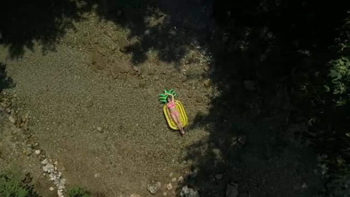 空中慢动作: 年轻的女性在菠萝漂浮物上晒黑，在河上滑行。