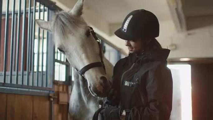 年轻的骑师女孩正在马stable里抚摸一匹白马。