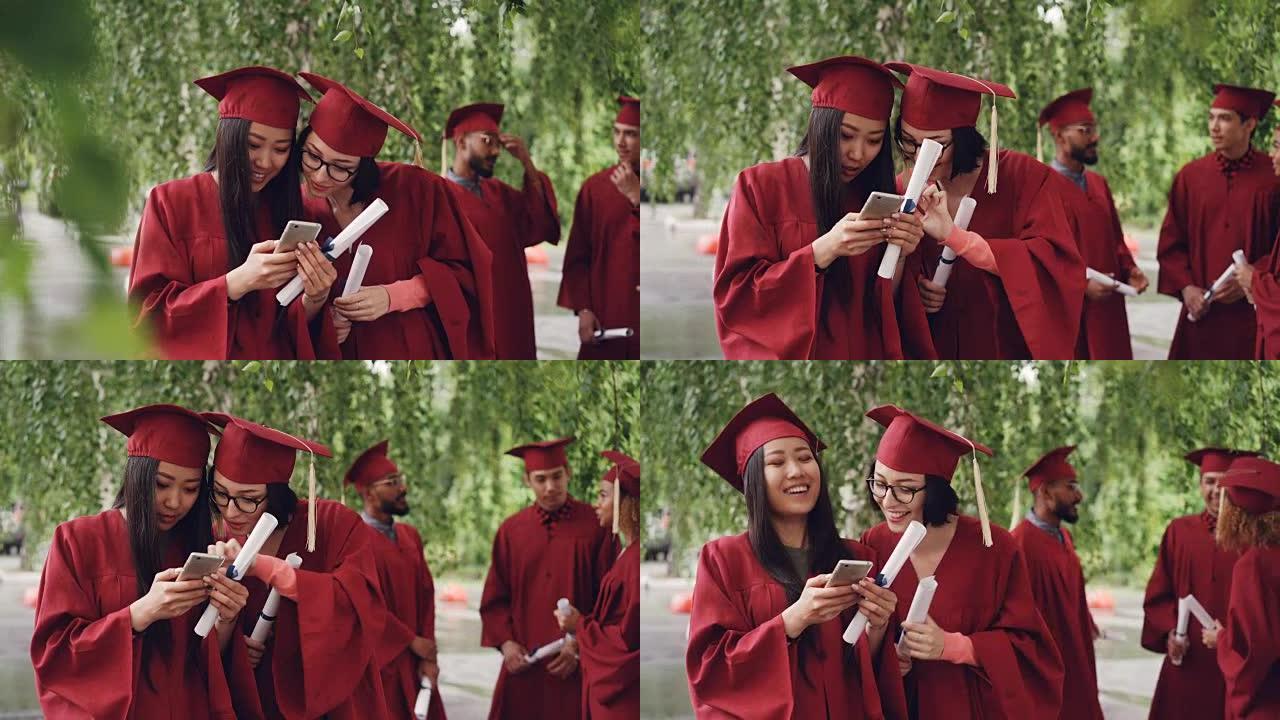 女毕业生正在使用智能手机看着屏幕上的说笑，站在户外拿着文凭，女孩穿着正式的礼服和帽子。
