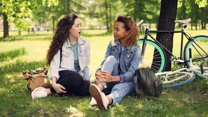 非裔美国女孩正在与坐在公园草坪上的白人朋友聊天，可见现代自行车和背包。混合种族友谊和对话概念。