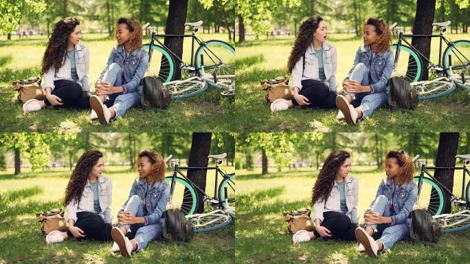 非裔美国女孩正在与坐在公园草坪上的白人朋友聊天，可见现代自行车和背包。混合种族友谊和对话概念。