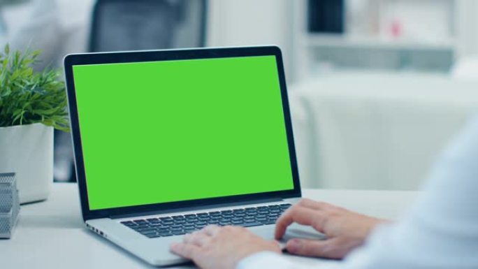 医生在绿屏打开的笔记本电脑上工作的特写镜头。他在后台的同事。办公室很现代，光线充足。