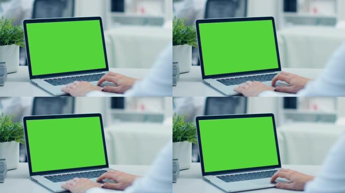 医生在绿屏打开的笔记本电脑上工作的特写镜头。他在后台的同事。办公室很现代，光线充足。